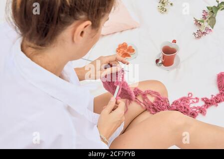 Grand angle de la femme douée se détendant sur un lit moelleux tout en créant des tricots pendant le travail à distance Banque D'Images