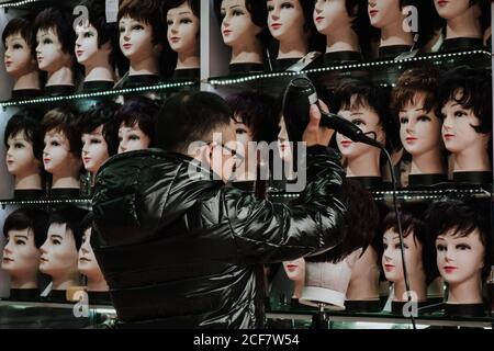 Shanghai, Chine - 12 décembre 2018 : vue arrière du vendeur de perruques mâles en veste chaude noire avec capuche, sèche-cheveux et coiffage des cheveux sur le mannequin tout en se tenant devant les tablettes avec mannequins sur le marché Banque D'Images