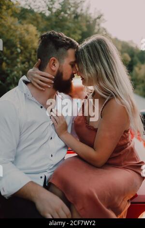 Homme à barbe tendre en chemise blanche et short noir enserrant femme souriante en robe tout en étant assis sur la capuche de voiture rouge sur l'autoroute près de la forêt verte Banque D'Images