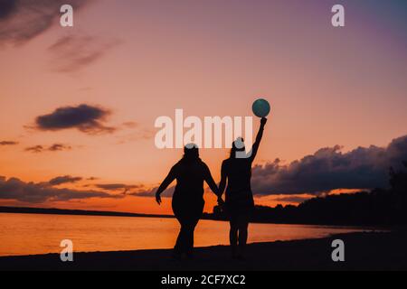 Vue arrière des femmes anonymes avec ballon tenant les mains et marche le long de la mer pendant le beau coucher de soleil Banque D'Images