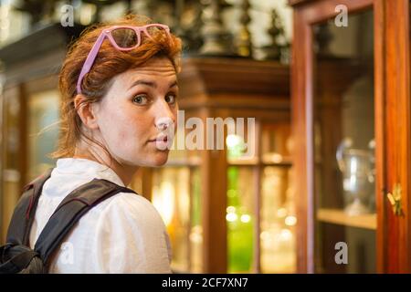 Vue latérale d'une jolie femme avec sac à dos regardant l'appareil photo au-dessus de l'épaule tout en se tenant sur un arrière-plan flou des vitrines dans la boutique de souvenirs au Sri Lanka Banque D'Images