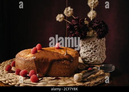 Appétissant gâteau fait maison avec des baies fraîches versées avec du chocolat garniture sur plateau en osier sur table avec fleurs Banque D'Images