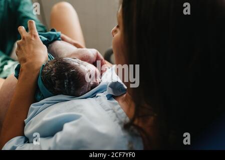 Grand angle de la gaie femme adulte embrassant le nouveau-né couvert de sang après avoir donné naissance dans la salle d'accouchement de l'hôpital contemporain Banque D'Images