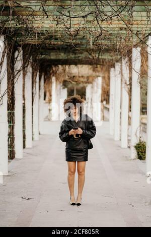 Jeune femme d'affaires en cuir noir rétro veste et jupe de navigation de smartphone tout en se tenant dans le passage voûté dans le parc avant le travail Banque D'Images