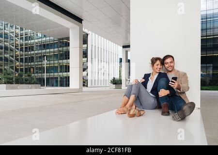 Couple joyeux s'embrassant tout en naviguant sur un smartphone assis à l'extérieur d'un bâtiment contemporain sur la rue de la ville ensemble Banque D'Images