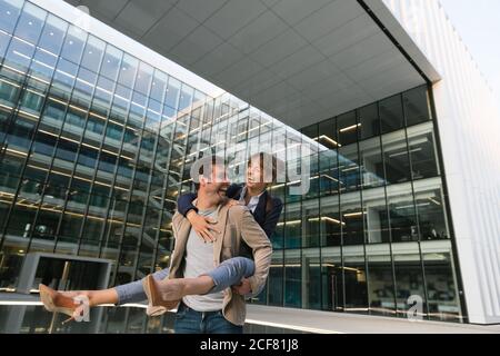 Un homme adulte heureux vêtue de vêtements décontractés porte une femme en train de se tenir debout et de rire dans la rue contre le centre d'affaires contemporain du centre-ville Banque D'Images