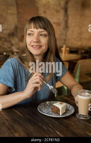 Blonde joyeuse femme avec des langes en bleu décontracté t-shirt tenir la cuillère avec un morceau de gâteau et s'asseoir à la table avec café et dessert Banque D'Images