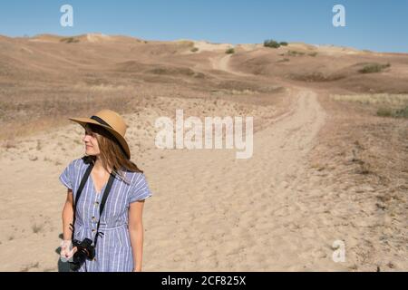 Femme adulte en chapeau de paille et robe avec caméra marchant sur le sentier de campagne Banque D'Images