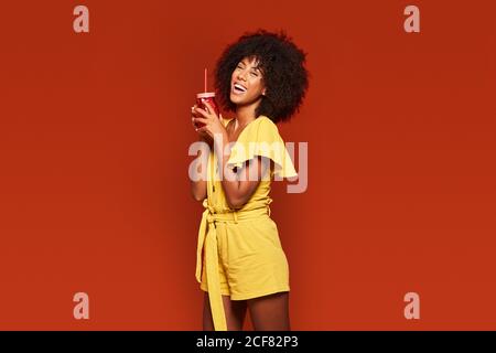 Jeune femme afro-américaine joyeuse aux cheveux bouclés tenant rouge bol avec paille et boisson sur fond rouge à l'appareil photo Banque D'Images