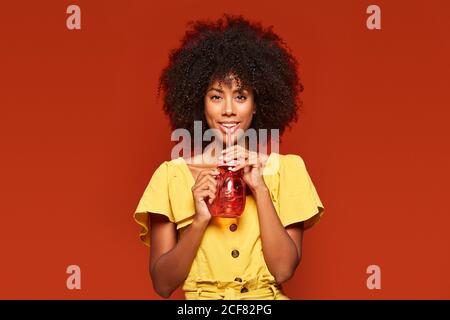 Jeune femme afro-américaine rêveuse aux cheveux bouclés qui tiennent rouge bol avec paille et boisson sur fond rouge à l'appareil photo Banque D'Images