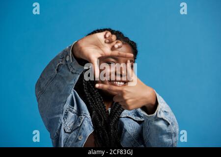 Bonne femme noire avec des tresses dans une veste en denim souriant et regarder l'appareil photo tout en cadrant le visage avec les mains contre le bleu arrière-plan Banque D'Images