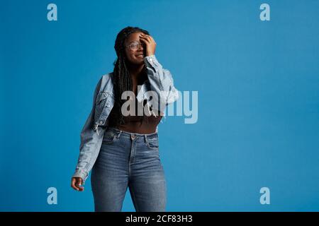 Femme noire de grande taille avec des tresses dans des vêtements décontractés souriant regarder la caméra couvrant la face avec la main contre un arrière-plan bleu Banque D'Images