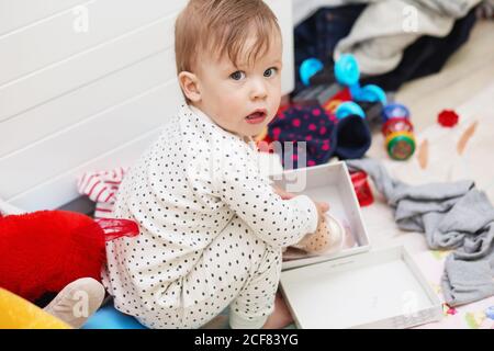 Mignon bébé fille en pyjama qui s'accroupette sur le sol essaie de mettez ses chaussures parmi les choses éparpillées et les jouets Banque D'Images