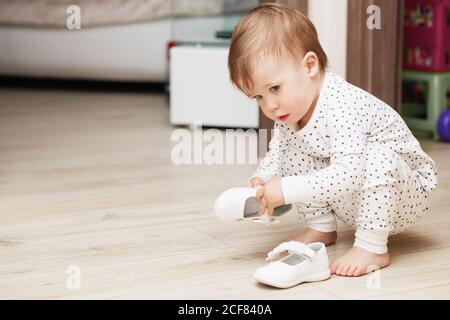 Mignon bébé fille en pyjama qui s'accroupette sur le sol essaie de mettez ses chaussures blanches dans la salle de jeux Banque D'Images