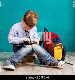 Un petit garçon joyeux avec un grand sac à dos tenant une tablette numérique sur fond bleu. Enfants jouant à des jeux sur un pavé tactile. Concept scolaire. Retour à Sho Banque D'Images