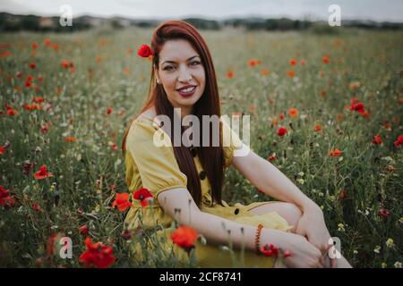 Femme adulte pensive à poil rouge, vêtu de robe jaune avec coquelicot rouge dans les cheveux et les lèvres rouges regardant sur l'épaule à l'appareil photo tout en étant assis seul dans un magnifique écrin de verdure avec des fleurs rouges et blanches contre des collines sous gris nuageux ciel Banque D'Images