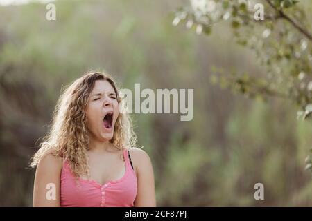 Femme endormie en tenue décontractée debout avec la bouche ouverte parmi arbres verts et bâillements pendant la journée Banque D'Images