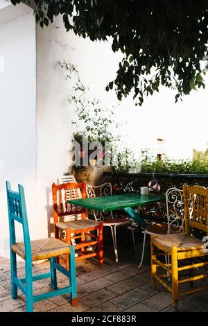 Ensemble coloré de table en bois avec différentes chaises placées à proximité clôture en métal avec plantes en pot couvertes de lierre sur la véranda de maison de campagne pendant la journée ensoleillée d'été Banque D'Images