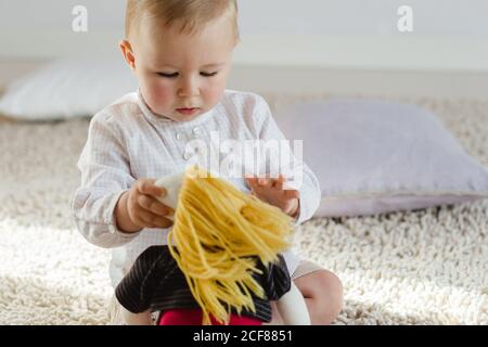 Mignon tout-petit dans des vêtements décontractés assis sur le sol et ayant amusez-vous avec une poupée douce Banque D'Images