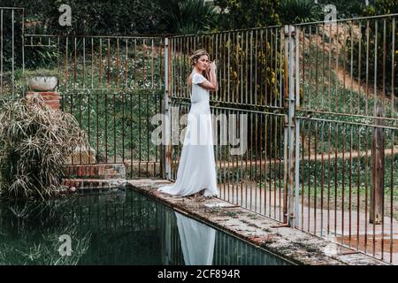 Vue latérale de la jeune femme en robe de mariée blanche se tenant sur la frontière avec les intempéries près de la clôture en métal et piscine calme dans le jardin Banque D'Images
