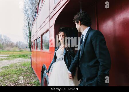 Mariée en robe de mariage et veste en cuir et marié costume élégant tenant les mains et se tenant près du double decker de merde bus en regardant les uns les autres Banque D'Images