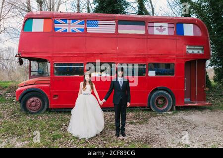 Mariée en robe de mariage et veste en cuir et marié costume élégant tenant les mains et se tenant près du double decker de merde bus en regardant les uns les autres Banque D'Images