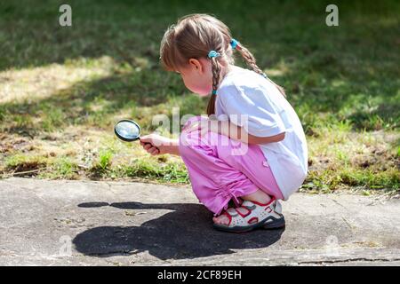 Petite fille curieuse à la recherche de quelque chose avec une loupe / loupe dans l'arrière-cour jeune enfant squatting, à la recherche de quelque chose sur le sol Banque D'Images