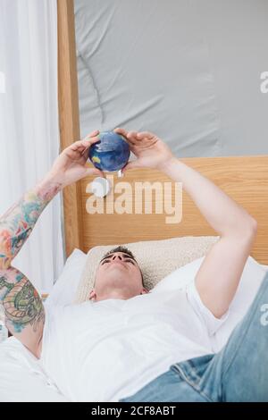 Beau homme tatoué en t-shirt blanc tournant dans les mains petites boule de terre tout en se relaxant sur le lit Banque D'Images