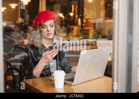 Jeune femme tendance dans le béret rouge sur le téléphone mobile pendant table avec ordinateur portable au restaurant Banque D'Images