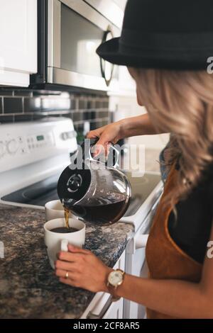 Vue latérale rognée d'une femme méconnue dans un chapeau avec montre-bracelet versez du café fraîchement préparé dans des tasses blanches en vous tenant debout cuisine à la maison Banque D'Images