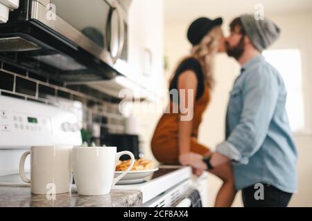 Vue latérale de l'homme en veste en denim et casquette à chaussettes embrasser petite amie dans un chapeau assis sur la table près des tasses avec boisson chaude dans la cuisine de l'appartement Banque D'Images