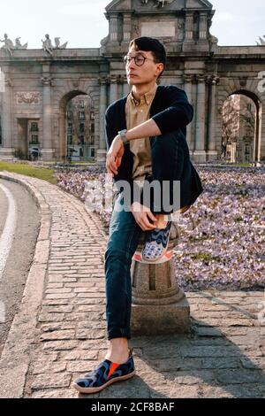 Corps complet de jeune asiatique tendance homme dans une tenue élégante et des lunettes assises sur une petite colonne de pierre posant et regardant loin de l'ancien beau bâtiment dans la rue Banque D'Images