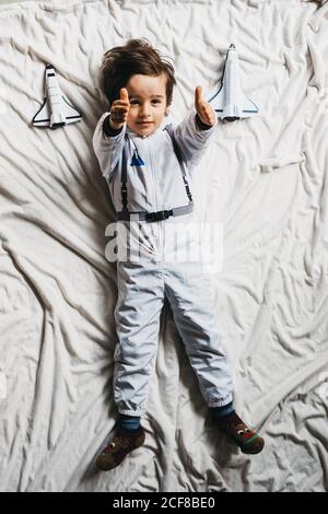 Vue de dessus d'un enfant souriant portant un espace allongé avec les bras surélevés sur le lit avec des vaisseaux spatiaux en peluche et regardant la caméra Banque D'Images