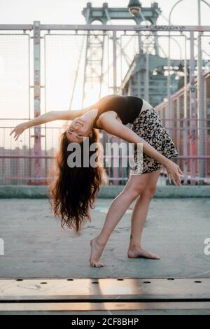 Vue latérale d'une jeune danseuse gracieuse en jupe décontractée effectuer une flexion arrière tout en se tenant pieds nus sur le pont à l'arrière allumé Banque D'Images