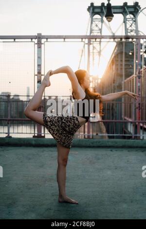 Vue latérale de la femme flexible dans une tenue décontractée pratiquant le yoga dans le seigneur de la danse poser tout en se tenant pieds nus dans le dos éclairé sur fond de paysage urbain et regardant loin Banque D'Images