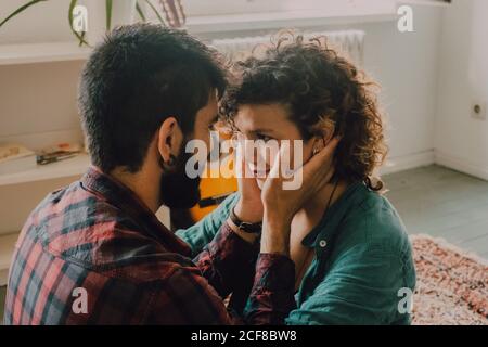 Vue latérale d'un couple tendre dans des vêtements décontractés en cuddling et souriant en vous asseyant sur la moquette au sol, à la maison les deux faces Banque D'Images