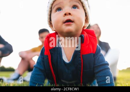 Mignon petit enfant ethnique dans des vêtements chauds souriant tout en rampant prairie verte pendant le week-end en famille à la campagne Banque D'Images