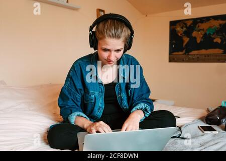 Joyeuse femme pensive dans le casque et les vêtements décontractés se concentrant sur l'écran est intéressant et vous pouvez utiliser votre netbook lorsque vous êtes assis avec vos jambes lit douillet croisé dans un appartement moderne et confortable Banque D'Images