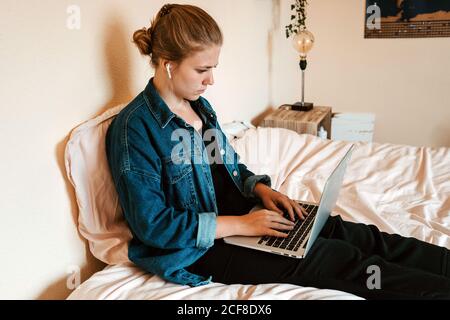 Vue latérale d'une femme bien pensée dans des écouteurs sans fil et des vêtements décontractés assis sur le lit avec un ordinateur portable contre beige mur dans l'appartement lumineux Banque D'Images