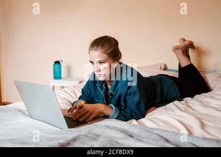 Femme réfléchie et concentrée dans des écouteurs sans fil et des vêtements décontractés couché sur le lit avec un ordinateur portable contre un mur beige appartement lumineux Banque D'Images