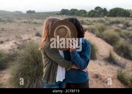 Homme et femme méconnaissables embrassant et cachant les visages derrière un chapeau tout en se reposant sur un fond flou du champ et du ciel de coucher de soleil dans la nature Banque D'Images