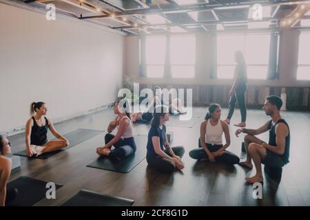 Grand angle de jeunes femmes et hommes divers dans les vêtements de sport assis dans lotus poser et avoir des discussions d'intérêt tout en se reposant après une formation de groupe dans un studio de yoga contemporain Banque D'Images