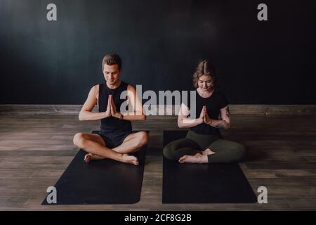 Calme jeune femme et homme dans les vêtements de sport avec les yeux fermés et les jambes croisées méditant ensemble tout en étant assis en pamasana position contre le mur arrière dans le studio de yoga contemporain Banque D'Images