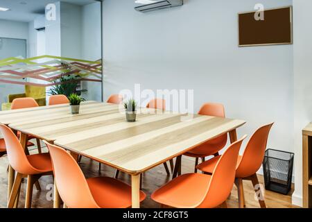 Intérieur simple et créatif dans une pièce contemporaine lumineuse et confortable de couleur orange chaises autour d'une grande table en bois contre des murs blancs et en verre Banque D'Images