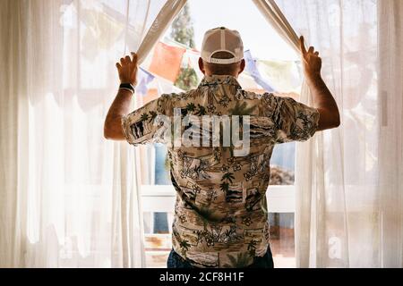 Vue arrière d'un homme méconnaissable en casquette et en ouverture de chemise rideaux de fenêtre et vue sur cour ensoleillée Banque D'Images