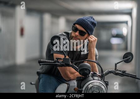 Homme barbu plein corps dans des vêtements décontractés et des lunettes de soleil dessus moto dans le couloir du garage moderne Banque D'Images