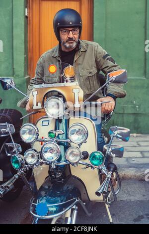 Homme mûr barbu dans un casque et des lunettes assis sur mesure moto avec beaucoup de phares sur la rue de ville Banque D'Images