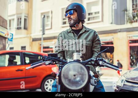 Homme barbu dans des lunettes de soleil et un casque assis sur une moto pendant le trajet sur la rue de la ville en regardant loin Banque D'Images