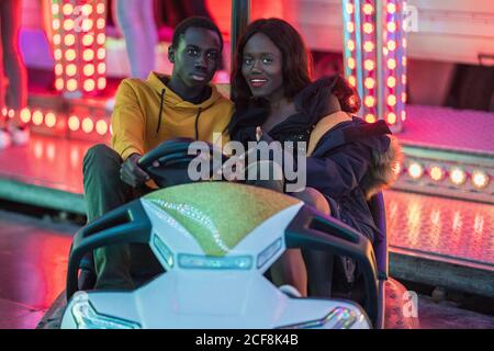 Homme et femme afro-américains souriant et roulant en voiture de pare-chocs pendant la date de nuit sur le terrain d'exposition Banque D'Images