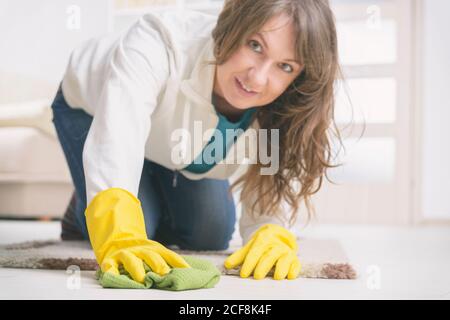 Femme souriante dans des gants en caoutchouc jaune essuyant la poussière sur le sol à la maison Banque D'Images